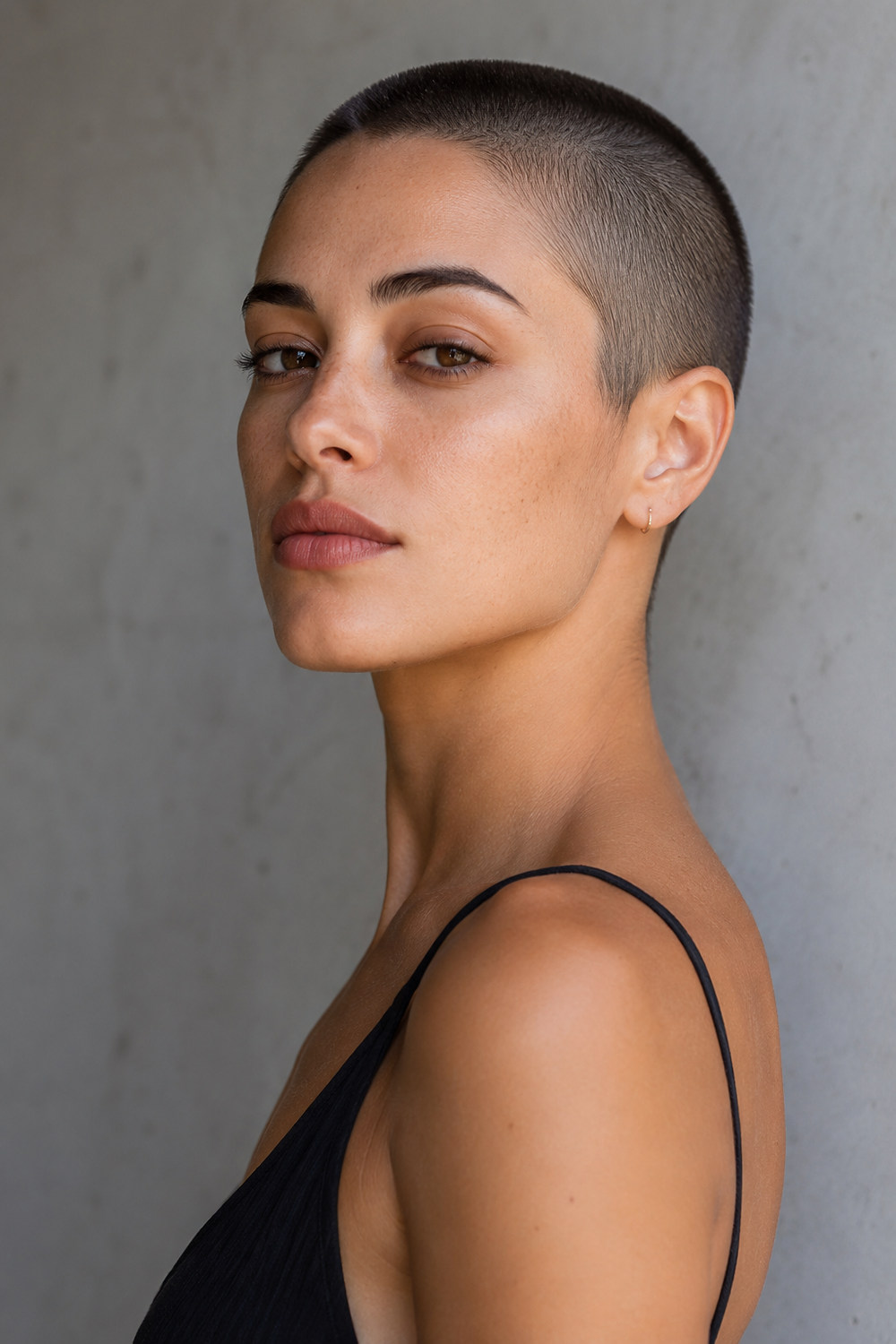 A woman with an even close buzz cut and strong clean silhouette against a neutral wall.